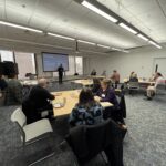 people sitting at a table looking at a speaker presenting